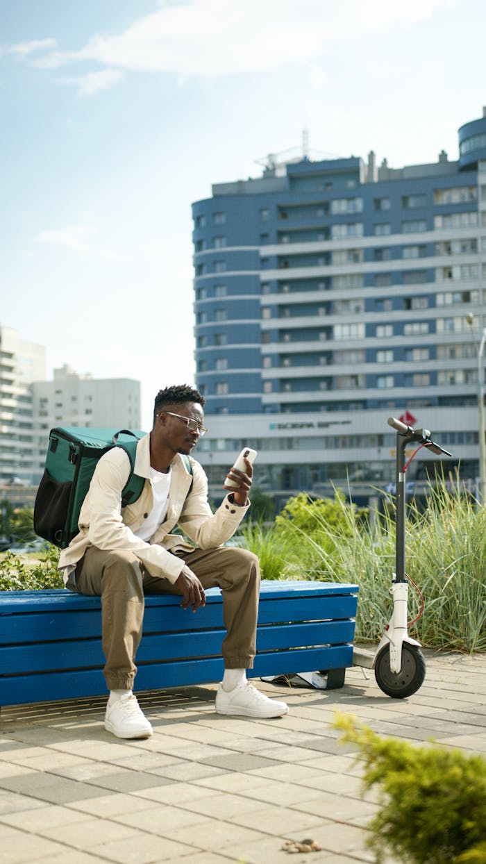 A man sits on a bench in the city, using his smartphone with an electric scooter nearby.